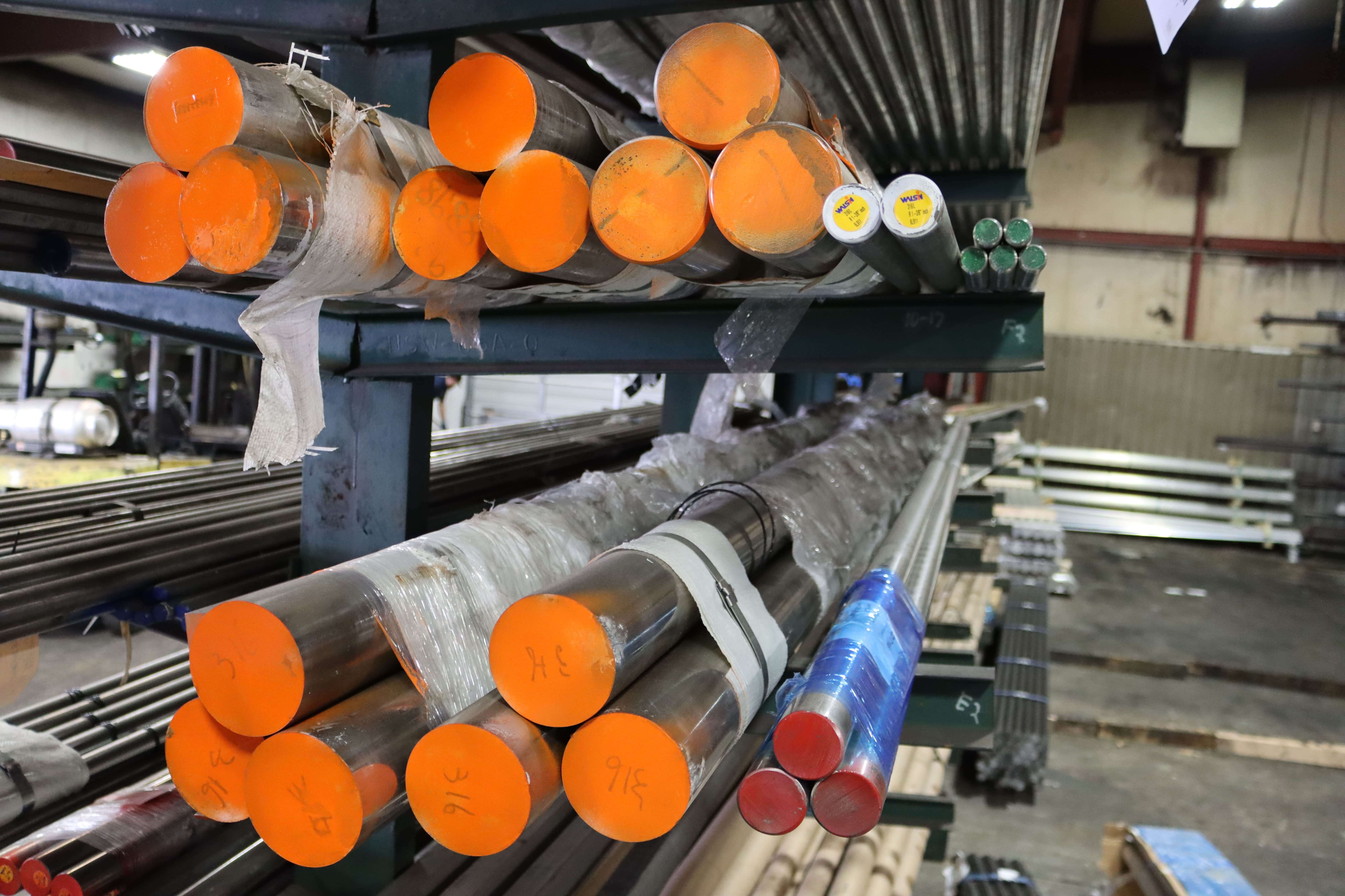 Storage rack with various metal rods wrapped in plastic, featuring bright orange and red caps in an industrial warehouse.