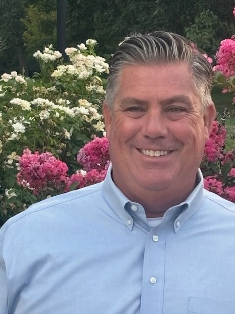 Portrait of Mike Hazel smiling in front of vibrant pink and white flowers.