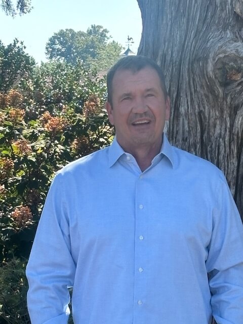 Portrait of Steve Reibert standing outdoors in front of greenery and a large tree.