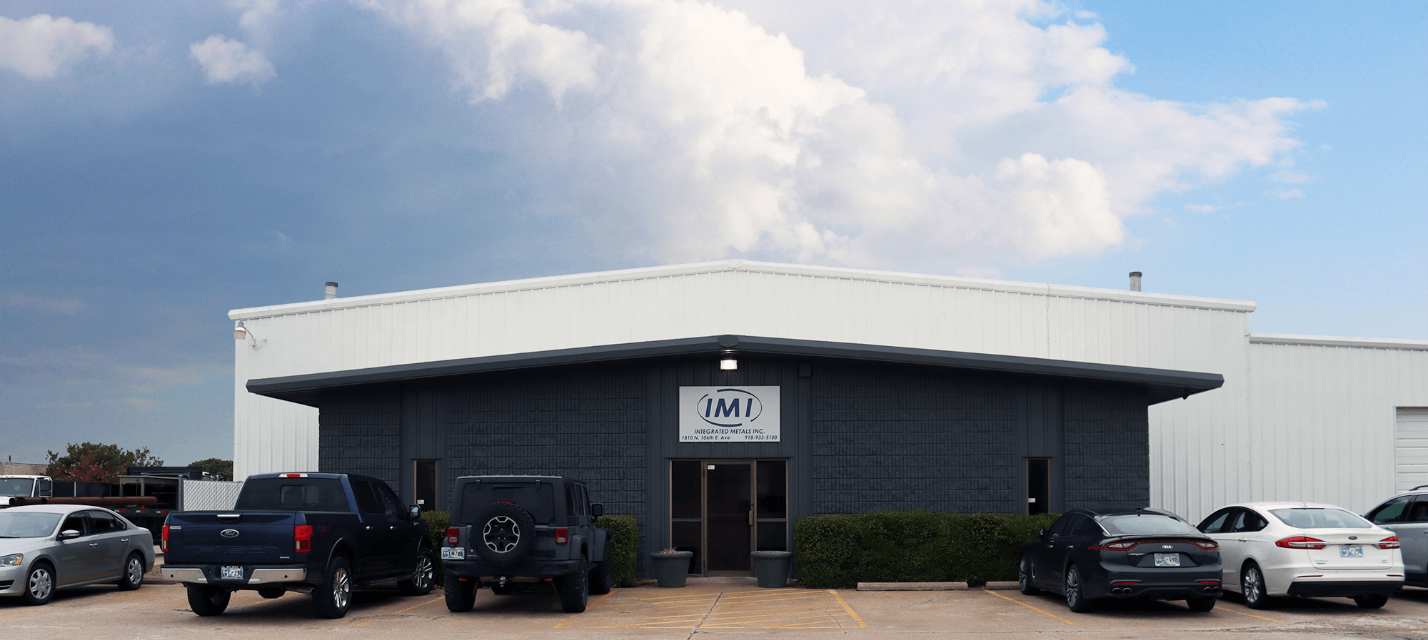 Front view of the Integrated Metals Inc. (IMI) building with a parking lot, cars, and cloudy sky overhead